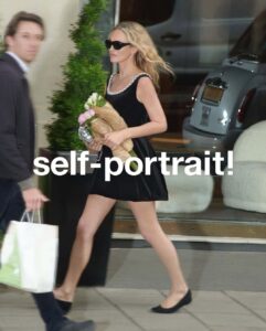 A woman in a black dress and sunglasses carries a bouquet and a small clutch while walking outdoors. Her hair flows naturally, and she exudes a confident, stylish demeanor. A blurred man walks past her on the sidewalk, holding shopping bags. Behind her, a parked car and greenery suggest an urban setting, likely a city street.