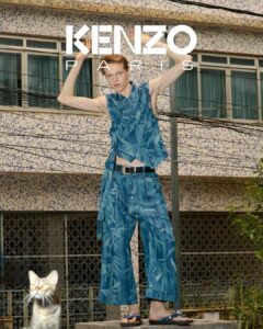 A person stands confidently on a raised surface, arms raised and showcasing a bold, patterned blue outfit featuring loose trousers and a sleeveless top. A cat is seen below, partially blurred. The backdrop is a building with small square tiles and several windows. Overhead, utility lines intersect, adding a dynamic urban feel to the scene.