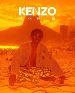 A person sits on a sandy beach with legs crossed, wearing patterned pants and a light, open-knit sweater. The background features a mountainous landscape with high-rise buildings, set against a vividly warm, orange sky that suggests sunset. The scene conveys a relaxed, summery vibe, with several other beachgoers visible in the distance, enjoying the scenic view.