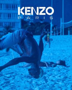 A person is mid-air, performing an impressive flip on a brightly illuminated beach. The setting is vivid with a strong blue tint. Behind, two people are lounging on the sand. The scene conveys a sense of energy and leisure, while nearby, a tall building and hints of distant vegetation complete the background.