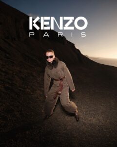 A person wears a stylish, brown outfit with faux fur details and a wide belt. The individual sports sunglasses and stands on a rocky terrain, creating a dramatic contrast with the dark backdrop. The lighting suggests dawn or dusk, casting a warm glow on the scene, while the expansive view hints at a rugged, natural landscape.