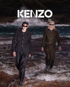 Two individuals walk confidently through the ocean shallows, dressed in stylish, dark suits. Their attire contrasts with the dynamic backdrop of crashing waves. Both wear sunglasses, adding an aura of modern coolness. The setting sun enhances the dramatic ambiance of the scene, casting shadows and highlighting their sleek outfits as they stride purposefully through the water.