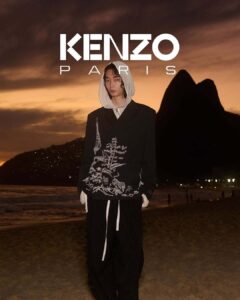 A person stands on a beach during sunset, wearing a dark jacket with intricate white embroidery and a hoodie. The sky is painted in warm hues, with dramatic clouds and two prominent silhouettes of mountains in the background. City lights twinkle faintly across the water, while the sandy shoreline stretches into the distance, adding a serene atmosphere.