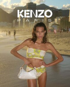 A person stands confidently on a beach, wearing a floral-patterned two-piece outfit while holding a white handbag. The sun casts a warm glow on the sandy shore and the distant cityscape, where tall buildings rise against a backdrop of misty mountains. The scene captures a blend of nature and urban life, creating a serene yet vibrant atmosphere.