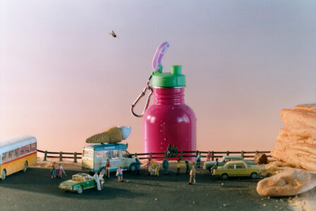 A vibrant scene features a large pink container with a green lid situated on a road, surrounded by miniature figures. People are gathered around, engaging in various activities near small vehicles, including an ice cream van. An array of figures, both standing and seated, adds life to the scene. A bird flies overhead against a calm sky.