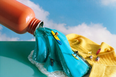 A vibrant orange container pours a wave of thick blue liquid over a textured surface. Miniature figures ride the wave on surfboards, depicting a playful surfing scene. A bright yellow fabric lies nearby, with smaller figures lounging, adding a whimsical touch. The backdrop features a pale blue sky with wispy clouds, enhancing the surreal atmosphere.