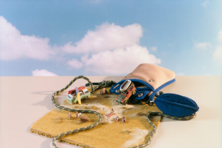 A beige pouch spills onto a yellow cloth resembling a desert scene. Miniature vehicles and tiny figures engage in a beach-like setting, complete with a surfboard and camping chairs. A knotted rope winds through the scene, while a blue flap from the pouch adds a splash of color. Fluffy clouds float in the background.