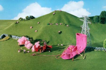 A whimsical scene features an idyllic green hillside populated with tiny pink sheep. In the foreground, miniature figures operate farm equipment that transforms the wool into vibrant pink yarn. This yarn is used to knit a massive sock, which is hung from a towering pylon. Playful and surreal, the landscape feels both pastoral and fantastical.