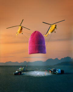 A large, pink knit hat is suspended in the sky by two yellow helicopters against a sunset background. Below, several miniature vehicles and figures are placed around a marked circle on the ground. The setting is a barren landscape with distant mountains under a gradient orange sky, creating a whimsical and surreal scene.