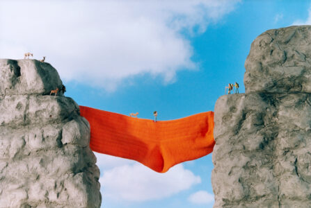 Two rocky cliffs stand opposite each other, connected by a large, vibrant orange cloth suspended like a bridge. Tiny figures, including people and animals, navigate the scene with a bright blue sky and fluffy clouds as the backdrop. The whimsical and imaginative setup creates a surreal landscape filled with a sense of adventure and creativity.