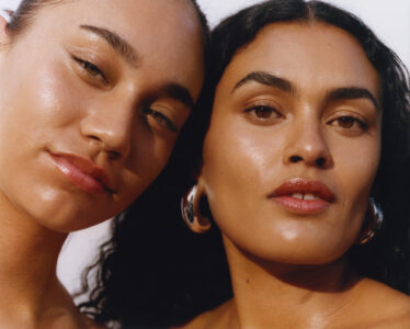 Two individuals are closely positioned, both with smooth, glowing skin and understated makeup. The person on the left has slightly closed eyes and a relaxed expression, while the individual on the right looks forward confidently with larger silver hoop earrings. The lighting highlights their facial features, casting soft shadows, and there is a sense of warmth and intimacy between them.