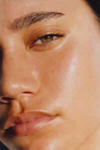 A close-up of a person's face highlights smooth, radiant skin with a natural glow. The focus is on one eye with lush lashes, shaped eyebrow, and soft, full lips. The complexion displays a healthy shine, with subtle freckles and fine skin details visible, creating a sense of freshness and vitality. The lighting accentuates facial features beautifully.