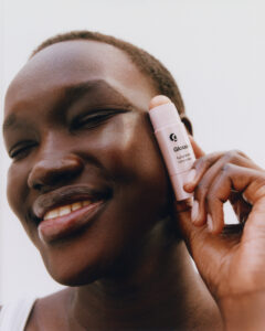 A person with a radiant smile gently holds a cylindrical skincare product to their cheek. The skin appears smooth and glowing, complementing the soft, neutral tone of the product casing. The background is minimal and clean, highlighting the subject's confident and joyful expression. The composition emphasizes natural beauty and a sense of contentment.