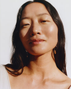 A close-up of a person with long dark hair and radiant sunlit skin. The individual has a calm expression, gazing slightly past the viewer. The natural lighting highlights their features, creating a warm and serene atmosphere. They wear subtle earrings and a simple top, contributing to an understated yet elegant appearance. The background is a soft, light color.