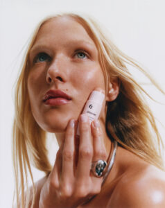 A person with long blonde hair gazes upwards, holding a small, light-colored cosmetic stick near their cheek. The face reflects a natural, dewy glow, complemented by soft pink lips. The individual wears a silver ring on their finger and a sleek silver necklace. The background is plain and bright, emphasizing the serene and minimalistic aesthetic.