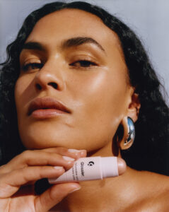 A person with smooth, glowing skin and sleek, dark hair gazes confidently forward. They wear a shiny, curved earring and hold a cosmetic product near their chin, showcasing its sleek design. The overall look conveys elegance and a focus on beauty, highlighting the luminous, well-highlighted texture of the skin and the sophisticated appeal of the accessory.