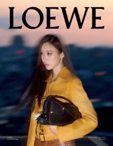 A person with long dark hair is seen wearing a mustard yellow jacket and brown pants. They hold a triangular black handbag with silver accents. The background features a blurred, dusk-lit cityscape in shades of blue and orange, with a hint of a sunset. The lighting creates a soft and moody contrast against the bright attire.