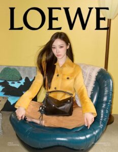 A woman sits cross legged in a deep teal leather chair, wearing a mustard jacket and tan trousers. A structured black handbag rests at her lap. Long dark hair falls smoothly over one shoulder. The room glows with warm yellow walls, patterned cushions, and soft studio lighting that sharpens textures and mood. Posture feels composed, confident, and slightly cinematic stillness.