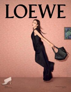 A person dressed in an elegant black outfit, holding a stylish black handbag, appears to be in motion against a patterned peach wall. A single white shoe is placed on a beige carpet. To the right, a small framed artwork hangs, adding a touch of color and sophistication to the scene.