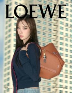 A young woman with long, dark hair poses confidently, wearing a dark sweater and light-colored jeans. She carries a large brown leather bag over her shoulder, featuring metallic accents. The backdrop showcases a tall building with numerous windows, suggesting an urban setting. Her expression is calm and poised, embodying a modern, stylish look.