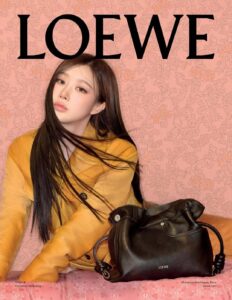 A person with long dark hair wears a mustard-colored coat, leaning slightly against a patterned backdrop. A black handbag with a smooth, structured design is prominently displayed in the foreground. The background features a vintage floral pattern in muted pink tones, adding an elegant touch to the overall composition. The scene conveys a sophisticated and stylish vibe.