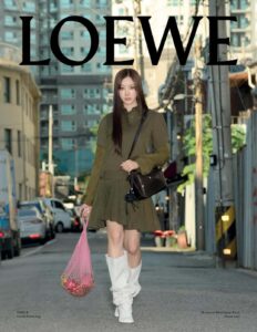 A woman with long hair stands in an urban setting. She's wearing a stylish olive green dress paired with tall white boots. Her right hand holds a pink net bag containing apples, and a dark handbag is slung across her body. The background features multi-story buildings, parked cars, and utility poles under bright daylight.