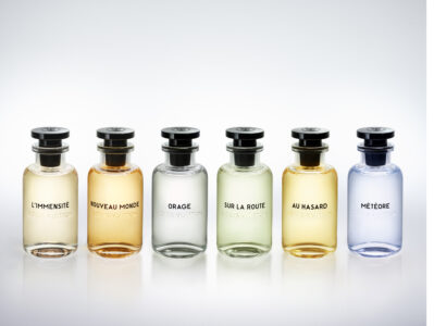 Six elegant glass bottles stand in a row, each differing slightly in color from light beige to pale blue. The bottles are identical in shape, with cylindrical bodies and sleek black caps, giving a modern yet classic appearance. Each container reveals a distinct liquid inside, creating a harmonious gradient across the arrangement. The setting is minimalist and sophisticated.