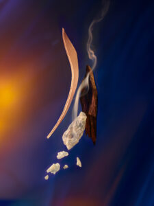 A composition of various organic elements appears set against a gradient background of deep blues and warm oranges. Delicate trails of smoke curl around a piece of dark wood, while nearby, translucent crystals cascade downward. A curved strip of material hovers nearby, its light tone accentuated by the surrounding shadows, creating a dynamic interplay of textures.