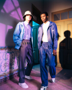 Two individuals stand casually in a stylish interior, each wearing blue-themed outfits. One wears a bucket hat and a light jacket, while the other sports a dark jacket and pants. Their shoes are white, complementing their outfits. The setting features decorative walls and a wooden door, with purple lighting casting dynamic shadows across the scene, adding artistic flair.