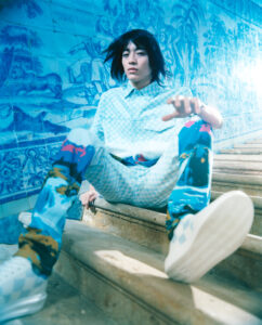 A person with medium-length dark hair is seated on stone steps, wearing a patterned outfit with a light blue shirt and colorful pants. The background features intricate blue and white artwork depicting people and animals. Sunlight enhances the vibrant hues and textures of the scene, creating a dynamic composition, blending classical and modern elements.