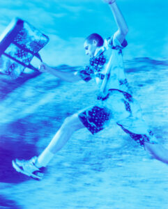 A person in motion leaps energetically against a dynamic, abstract blue background. They wear a vibrant, patterned outfit with matching shorts and shirt, paired with stylish sneakers. One arm is extended forward, holding a large, patterned bag. The overall feel is energetic and expressive, highlighting movement and style in a surreal, dreamlike setting.