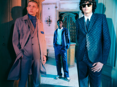 Three individuals are standing in a stylish setting. The first person on the left wears a long coat and high-collared shirt, exuding sophistication. The middle figure stands slightly further back, dressed in a casual blue jacket and pants. The person on the right sports a textured suit with sunglasses, adding a cool, modern vibe to the scene.