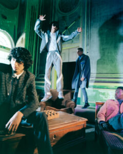 A group of four individuals is gathered in a vintage-looking room with ornate walls and furniture. One person energetically stands on a chair, arms raised, while others sit or stand nearby, wearing stylish, modern clothing. The lighting creates a dramatic and dynamic atmosphere, enhancing the contrast between the historical setting and contemporary fashion.