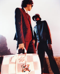 Two individuals, styled in fashionable attire, stand near a rocky coastline. The scene is imbued with a warm, artistic glow. One holds a large, patterned bag, suggesting elegance and luxury. The person on the left wears a dark, tailored suit, while the other sports a darker jacket and hat, creating a striking contrast in the composition.