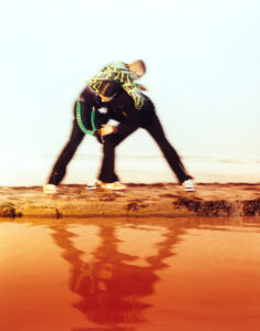 Two individuals engage in a playful embrace, their blurred forms creating a dynamic sense of movement. The sandy ground beneath them borders a vivid, reflective pool of orange-hued water. The scene conveys a lively, energetic atmosphere, with one person's clothing featuring a distinctive green pattern that contrasts with the surroundings. The background is minimal, highlighting the interaction.