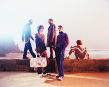 Five fashionably dressed individuals stand and sit on a concrete barrier near a serene beach. The group exudes a mix of casual and elegant styles, with vibrant and contrasting outfits. Two of them stand prominently in the foreground, while the others are positioned slightly further back, one seated and a couple standing. The background contains a soft, warm glow, adding a dreamy atmosphere.