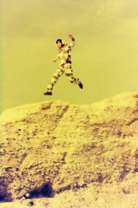A person wearing a colorful patterned outfit is captured mid-air, seemingly leaping off a rocky terrain. The background features a muted yellow sky, giving the scene an aged, vintage feel. The rocky surface appears textured with rough, uneven details, contrasting the smooth, pale sky. The individual's dynamic pose conveys a sense of motion and freedom.
