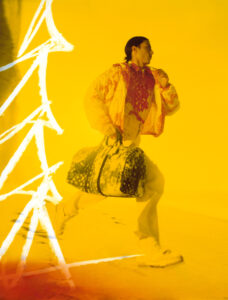 A person moves dynamically, dressed in a vibrant, patterned outfit comprising a jacket and trousers. They carry a large, stylish duffel bag with distinctive graphics. The scene is enveloped in warm yellow tones, creating a sense of movement and energy, accentuated by striking white streaks that suggest motion or light effects. The atmosphere feels bold and modern.