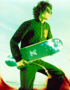 A person with curly hair and sunglasses holds a skateboard, surrounded by a dreamy, yellowish-green hue. They wear a patterned jacket and dark pants, with a keychain hanging by their side. The background features a light blue sky with soft clouds, creating a relaxed, stylish atmosphere suggestive of a sunny day by the shore.