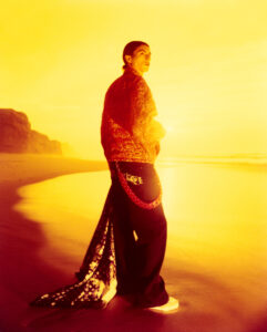 A person stands on a sunlit beach, casting a long shadow on the sand. Cliffs are visible in the background under a vibrant yellow-orange sky. The individual is dressed in a textured red jacket and dark trousers with a decorative chain and scarf, creating a colorful, dramatic contrast against the glowing, serene coastal setting.