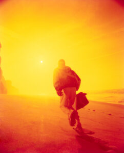 A lone figure, wrapped in vibrant hues of orange and yellow, walks along a sandy beach. The sun's warm glow creates a surreal atmosphere, casting long shadows and illuminating the path. Waves gently approach the shore, while the person carries a bag, striding towards the horizon, embodying a sense of journey and tranquility amidst the radiant light.