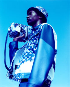 A person stands against a vivid blue backdrop, wearing a stylish outfit featuring vibrant patterns and textures. They hold a patterned bag over their shoulder. The attire includes a jacket with mixed materials and a matching hat. The overall composition emphasizes a modern, fashionable look, emphasizing bold colors and sleek design in a striking perspective.