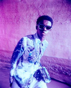 A stylish individual dons oversized sunglasses and a long-sleeve shirt adorned with intricate patterns, featuring hues of blue and white. A matching crossbody bag hangs from the shoulder. The background showcases a textured wall illuminated with vibrant gradients of purple and pink, creating an artistic, high-fashion ambiance with a modern, vibrant twist.