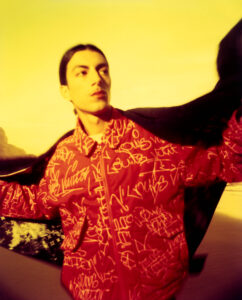 A person with long hair stands against a warm, yellow background. They wear a red jacket adorned with white graffiti-like patterns. The light creates a smooth, glowing effect, casting soft shadows. The individual holds a dark fabric or cloth that billows behind them, adding a sense of motion and drama to the scene.