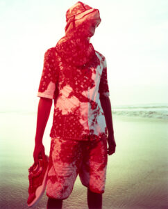 A person stands on a tranquil beach, dressed in a coordinated outfit with a vibrant red tie-dye pattern. They wear a head covering and hold matching red sneakers in one hand. The soft lighting of the scene creates a serene atmosphere, while the gentle waves of the ocean add an element of calm in the background.