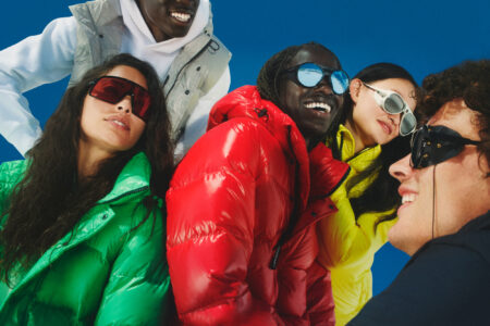 A group of people enjoys a sunny day against a vibrant blue background. They wear brightly colored puffer jackets in red, green, and yellow, and everyone sports various stylish sunglasses. The mood is cheerful, with smiles and laughter shared among them. Shadows and lighting suggest a dynamic, fun atmosphere, as they lean in close, highlighting friendship and joy.
