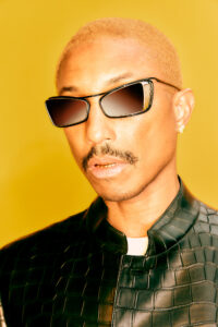 A person with short, bleached hair sports angular sunglasses and a subtle mustache. They wear a dark, textured jacket with a high collar. A stud earring gleams on one ear, and their lips reveal a hint of gold. The background is a vivid yellow, enhancing the overall warm, stylish vibe of the composition.