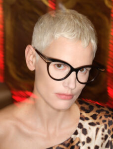 A person with short platinum blonde hair and large black-rimmed glasses gazes confidently forward. Their expression is composed, with softly pursed lips. They wear an off-the-shoulder garment featuring a bold leopard print pattern. The background is decorated with patterns and hints of red lighting, creating a warm and stylish atmosphere. The overall look is modern and sophisticated.