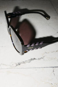 A pair of elegant brown sunglasses rests on a light marbled surface. The wide, square frames create a bold look, while intertwined purple accents along the arms add a touch of sophistication. The lenses reflect a subtle shine, capturing the surrounding light. Shadows cast gently around, enhancing the striking design of the eyewear with a modern flair.