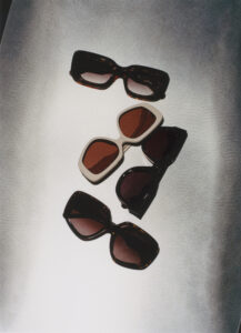 Four pairs of sunglasses are displayed on a textured surface. Two have dark frames with brown lenses, one has a light frame with a retro design, and the last one features a sleek, modern look. The arrangement forms a relaxed, casual vibe, highlighted by subtle lighting that casts gentle shadows, creating a sense of depth and style.